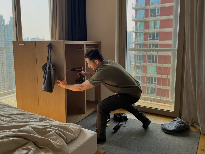 Technician reassembling a wooden wardrobe with a cordless drill in a bedroom with a skyline balcony